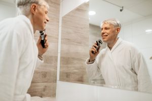 Cheerful adult man shaves at mirror in bathroom
